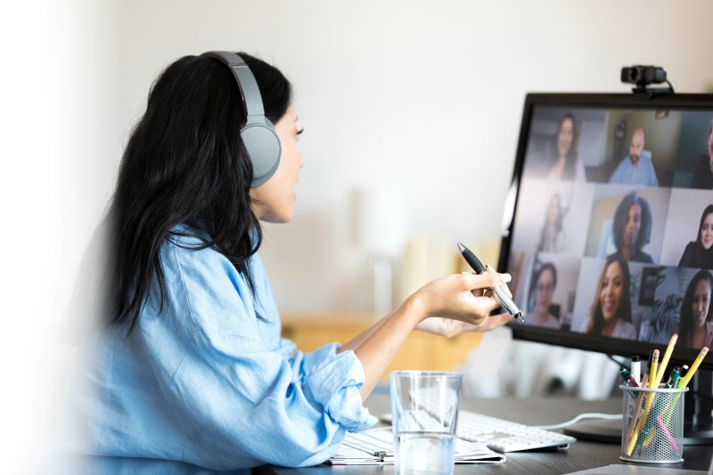 Kvinna i videomöte med flera personer, har hörlurar på sig och talar medan hon gestikulerar med en penna.